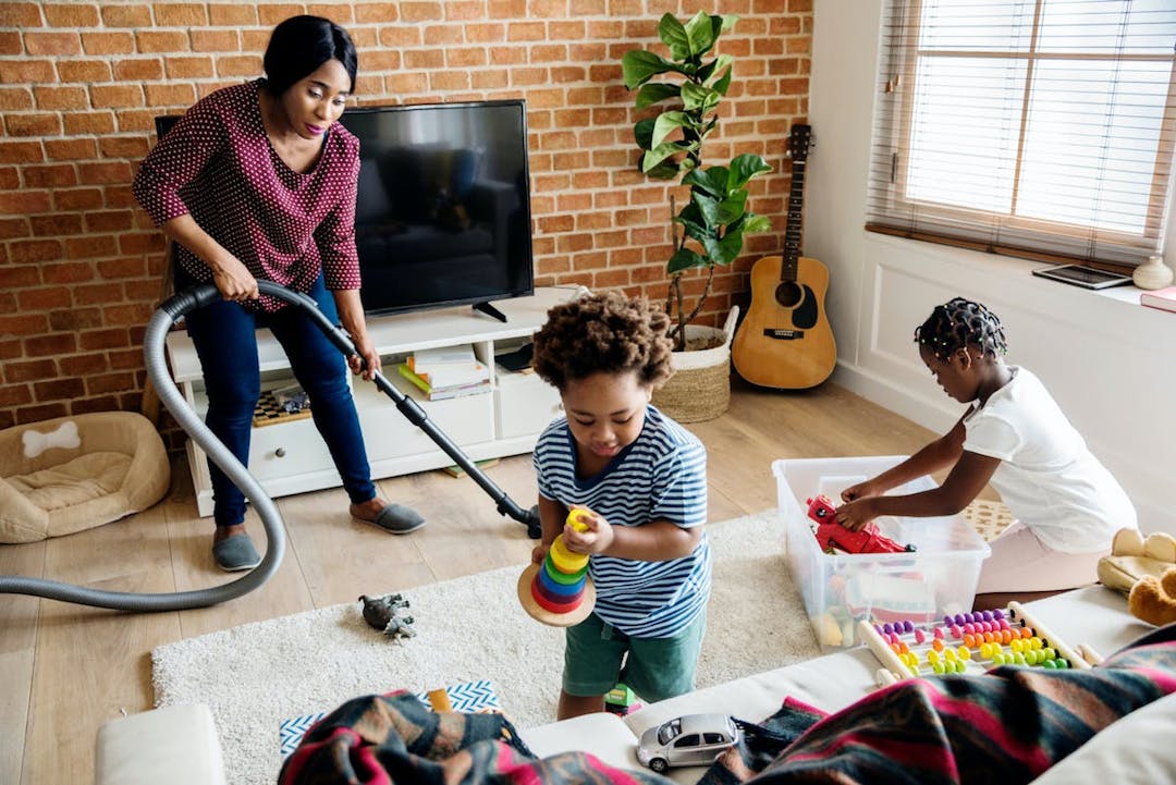 A woman vacuuming the floor while two kids play.